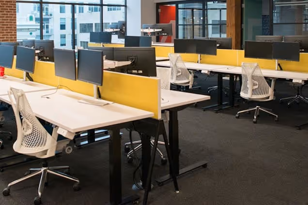 White chairs at desks with dual monitors and yellow privacy screens on a dark grey carpeted office floor.