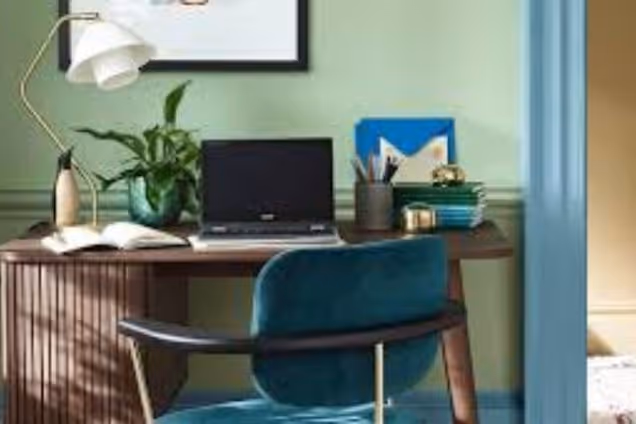 A laptop and lamp sit on a desk next to a chair in a room with green walls and a teal seat.