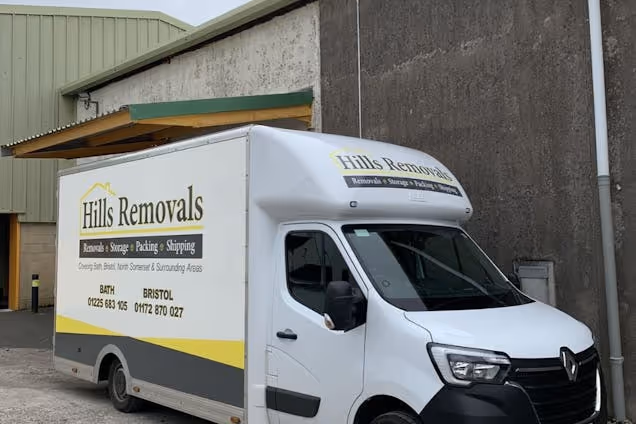 A white "Hills Removals" van with yellow accents parked in front of a warehouse and a stone wall.