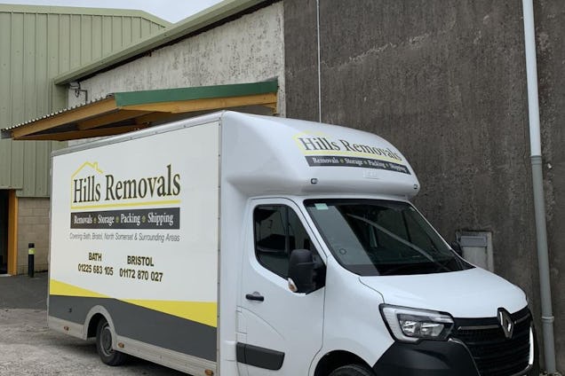 A white "Hills Removals" van with yellow accents parked in front of a warehouse and a stone wall.