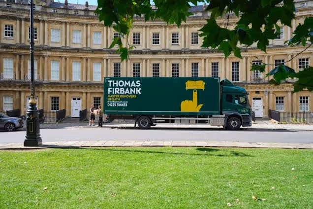 A green truck with a logo sits on a street in front of a row of buildings and a lawn.