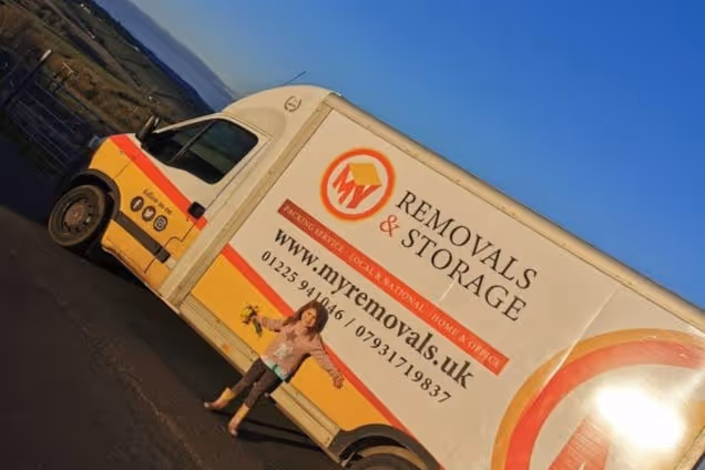 A white and orange removal van with a "Removals & Storage" logo on a road near a hillside under a blue sky.