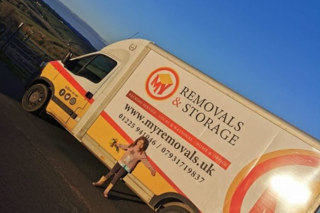 A white and orange removal van with a "Removals & Storage" logo on a road near a hillside under a blue sky.