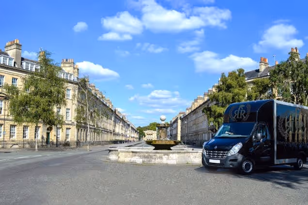 A black moving van parked on a wide street with stone buildings and a fountain under a blue sky.
