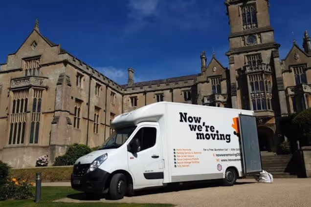 A white moving van with "Now We're Moving Van" on it parked on a road in front of a stone building with towers and a lawn.