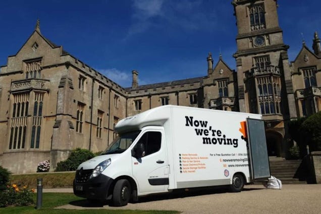 A white moving van with "Now We're Moving Van" on it parked on a road in front of a stone building with towers and a lawn.