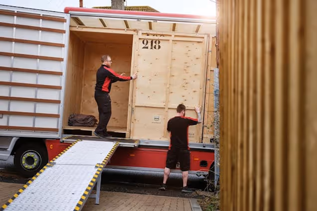 Two men loading a large wooden storage crate onto the back of a moving truck via a metal ramp.
