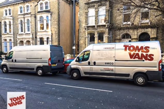 Two silver vans, one with a "Toms Vans" logo, parked on a street in front of stone houses.