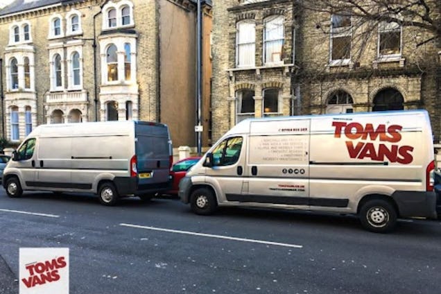 Two silver vans, one with a "Toms Vans" logo, parked on a street in front of stone houses.