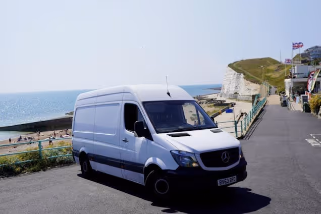 A white van parked on a coastal road overlooking a beach and white cliffs under a blue sky.