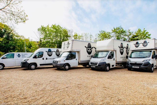 A fleet of five white removal vans and trucks parked on a gravel lot in front of trees.