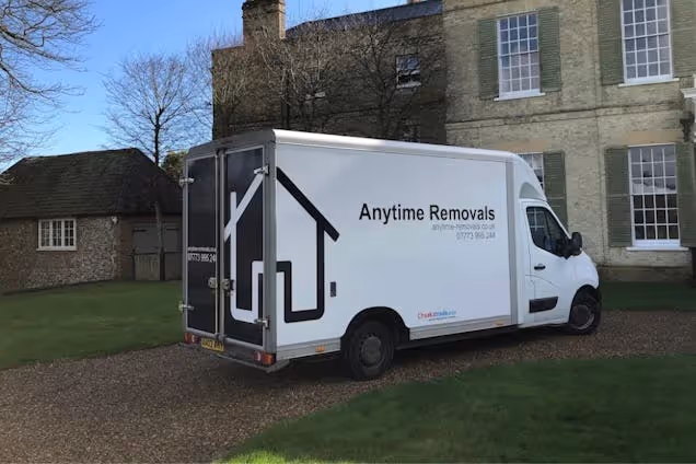 A white Anytime Removals truck parked in front of a stone house with a thatched roof cottage nearby.