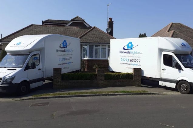 Two white removal vans with blue house logos parked on a street in front of a bungalow.