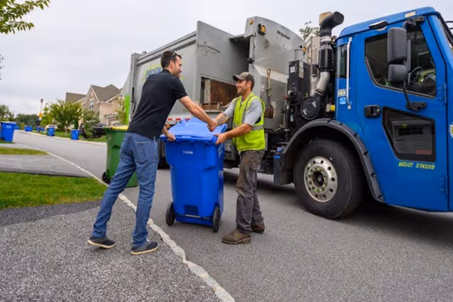 A man in a black shirt hands a blue recycling bin to a worker in a yellow vest next to a blue rubbish truck.