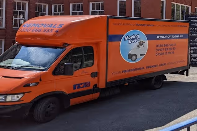 An orange Moving Ewe Ltd removal van parked on a street in front of a brick building.
