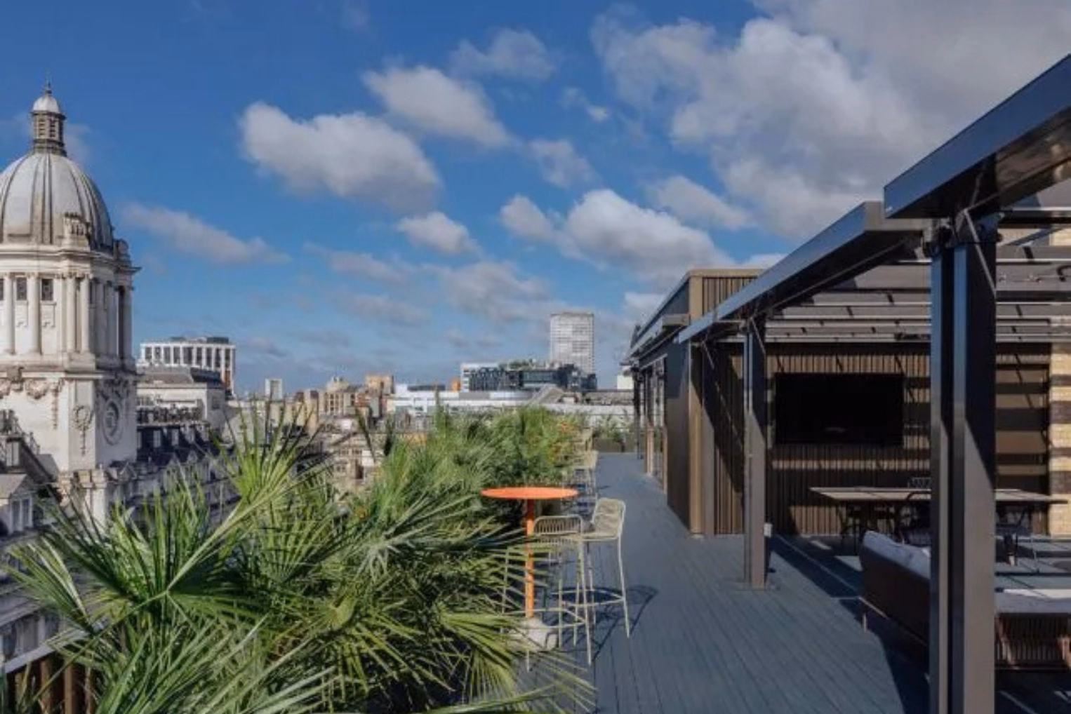 An orange table and green stools on a blue floor near plants. Greyfurniture stands under a black pergola near a dome and buildings under a sky.