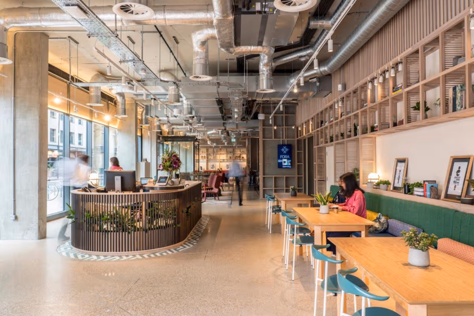 People sat at wooden tables on a green bench while a person walks past a wooden desk. Teal chairs, wood shelves, and silver pipes are in a room.