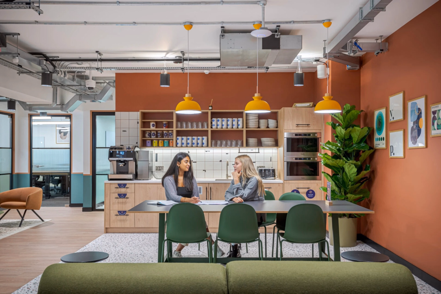 Two women sat at a black table with green chairs, looking at papers. A green bench and wood cabinets near an orange wall under yellow lights.