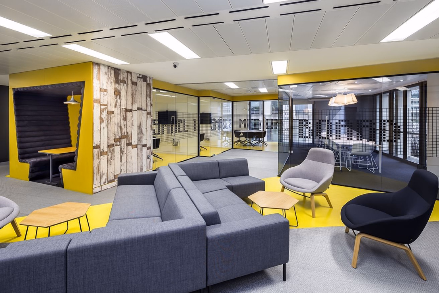 Grey sofa, grey armchair, and black armchair with wood tables on a grey and yellow floor near yellow walls and glass rooms.
