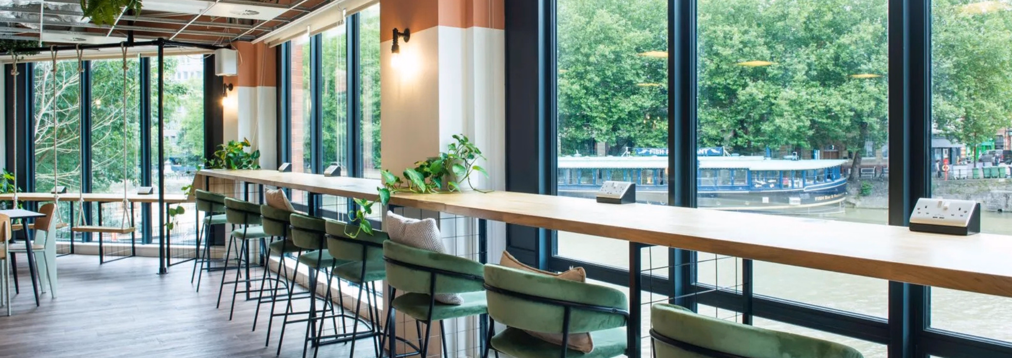 Green chairs and a swing at a wood counter near windows. A river, trees, and a boat are in the background.