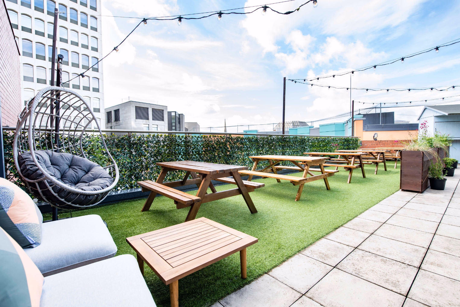 Wood picnic tables, a grey hanging chair, and grey seats with pillows sit on grass. String lights hang between buildings under a sky.
