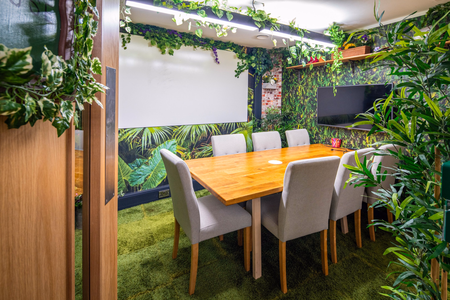 Grey chairs sit around a wood table on a green carpet. Plants, a screen, and a TV stand in a room with leaf graphics.
