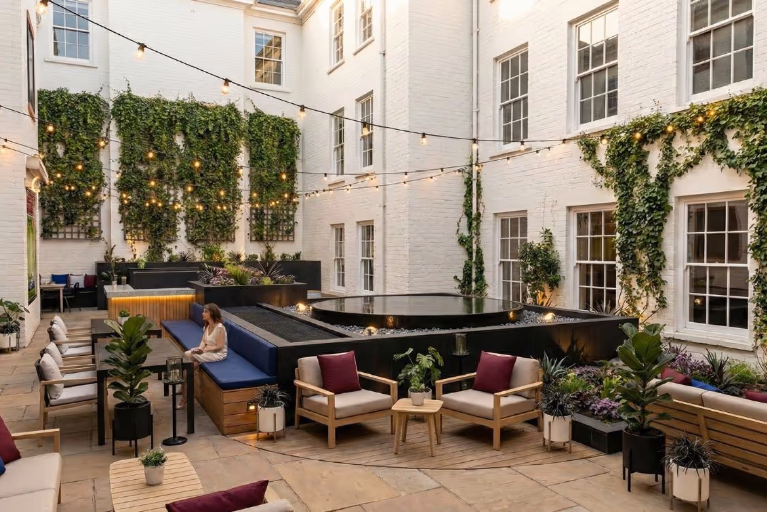 A woman sits on a blue bench. Tan armchairs with purple pillows and black tables sit in a courtyard with a fountain and white walls under lights.