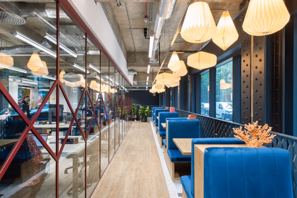 People work at desks behind a glass wall with red frames across from blue booths and tan tables. Yellow lights hang from a ceiling over a wood floor.