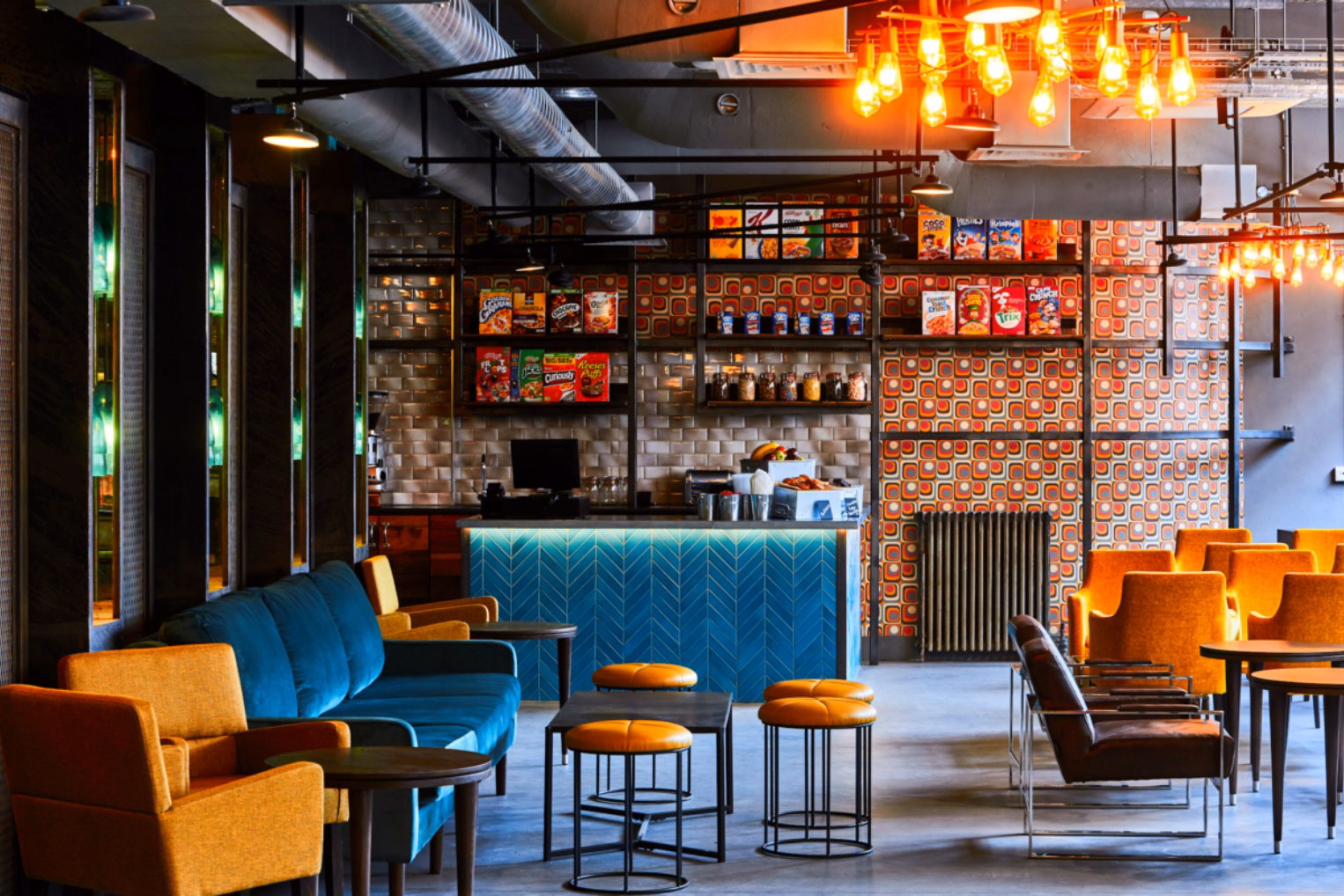 A blue sofa, yellow armchairs, and orange stools sit near black tables on a grey floor. A blue counter and shelves with cereal boxes stand under orange lights and silver pipes.