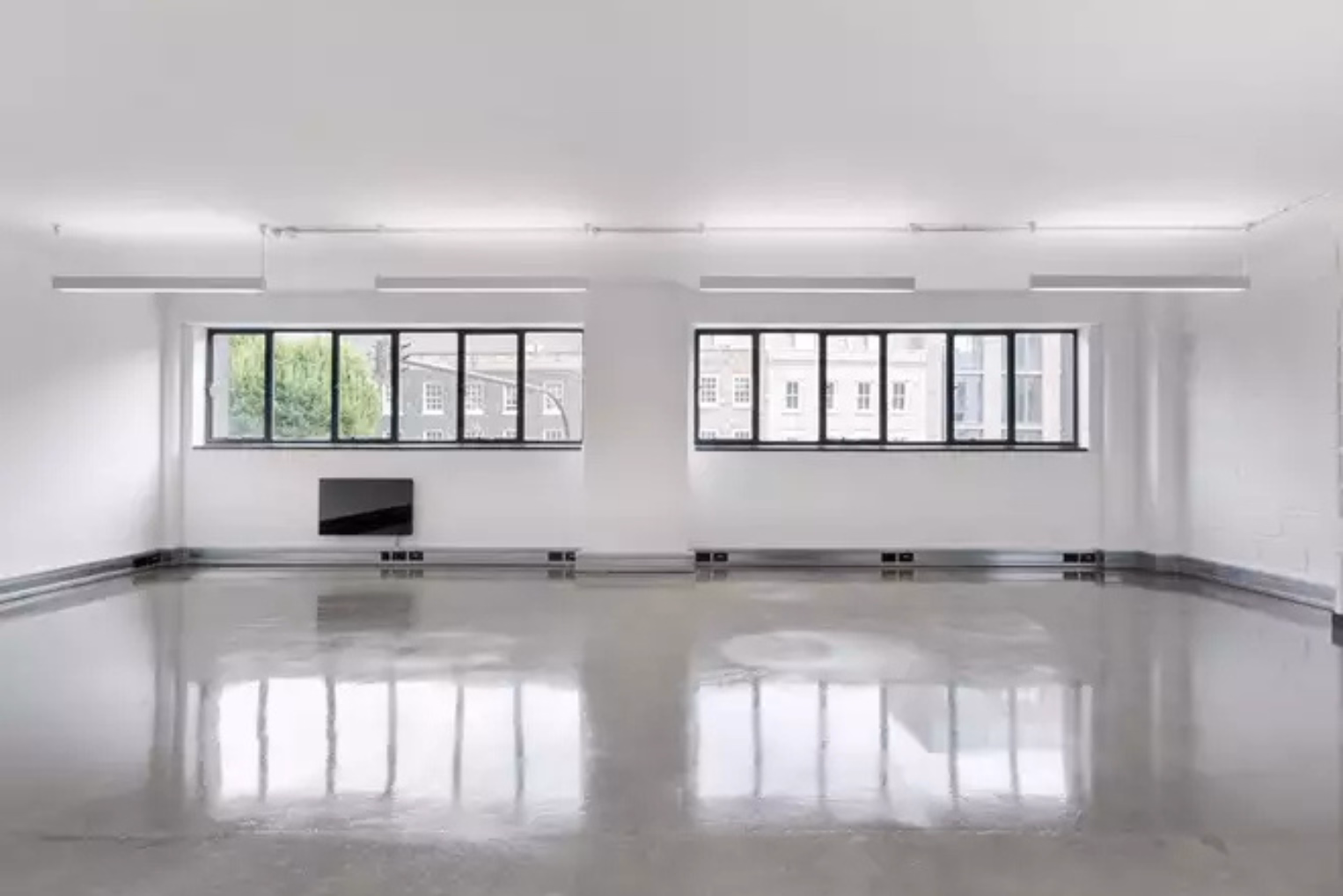 A grey floor stands under white walls and windows with black frames. White lights hang from a ceiling near a black panel.