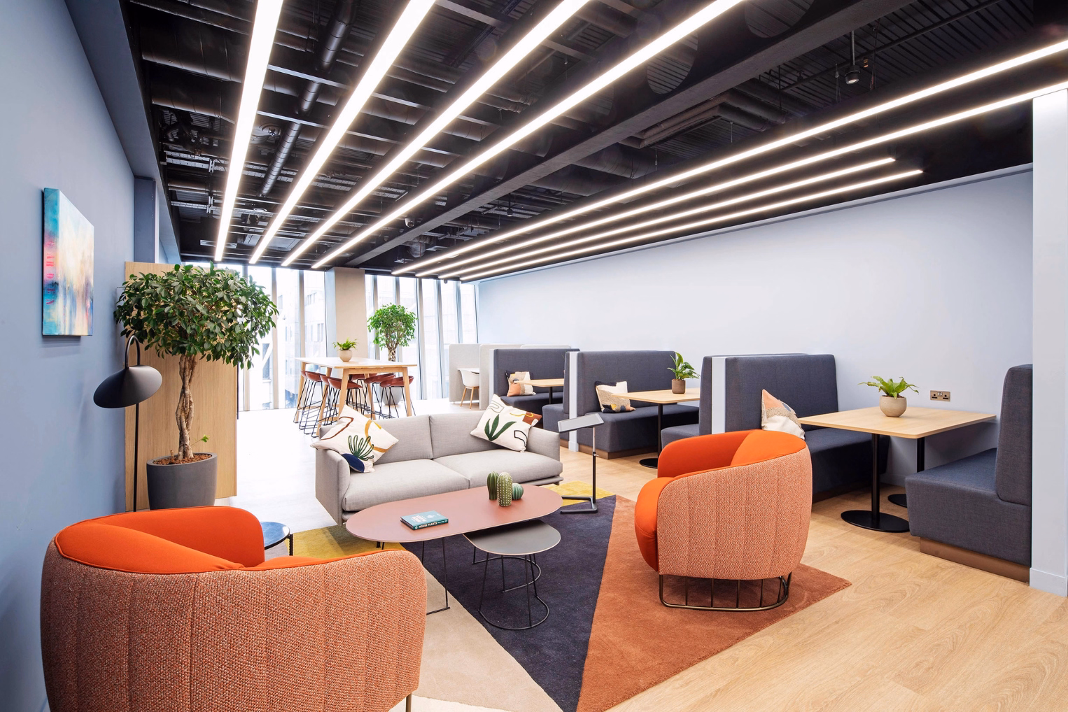 Orange armchairs, a grey sofa, and a brown table sit on a rug across from grey booths with tan tables. White lights hang from a black ceiling over a wood floor.