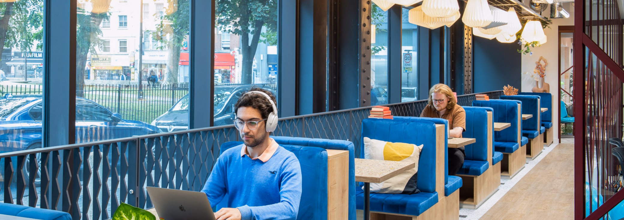 Men use laptops at tan tables in blue booths near windows. White lights hang from a ceiling over a wood floor.