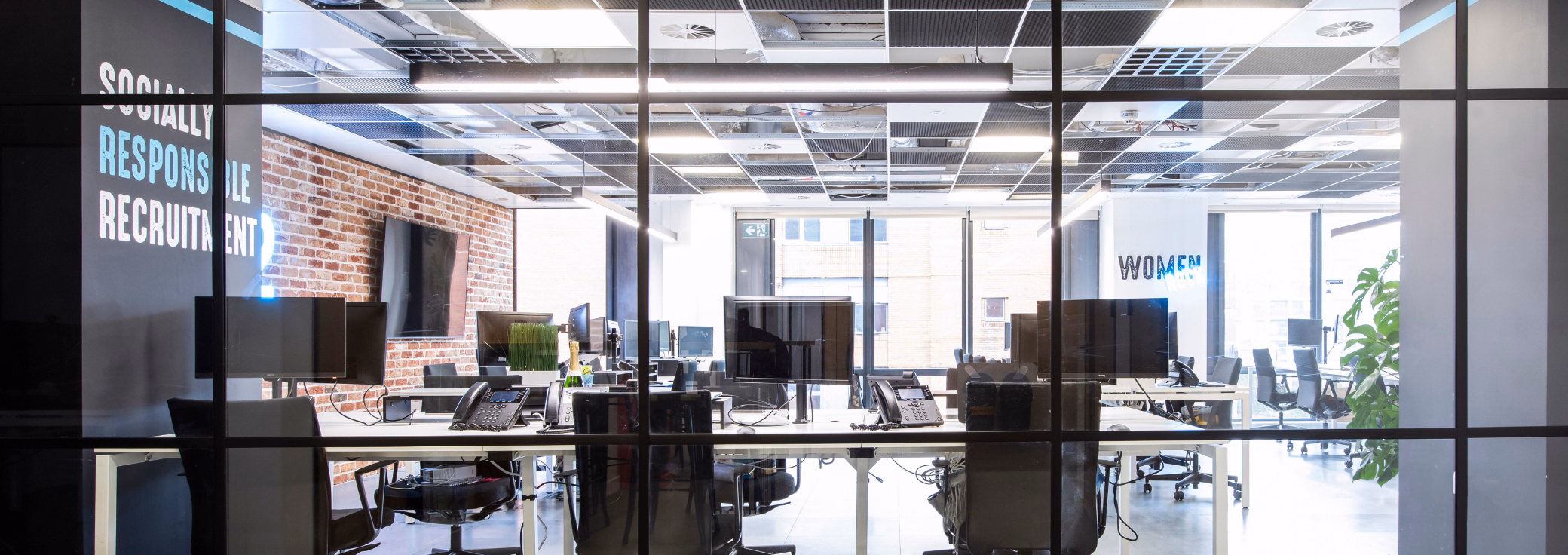 Black chairs and monitors sit at white desks behind a glass wall. A brick wall and lights are in the office.