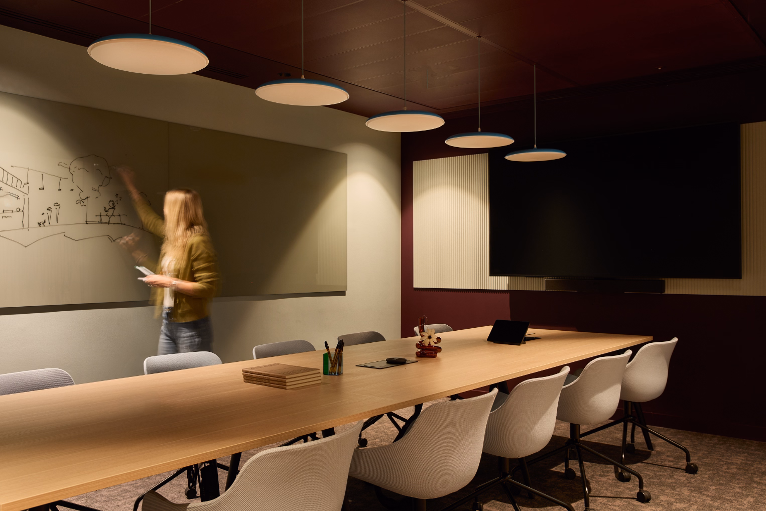 A woman writes on a whiteboard next to a wood table and white chairs. Lights hang from a ceiling near a black screen on a wall.