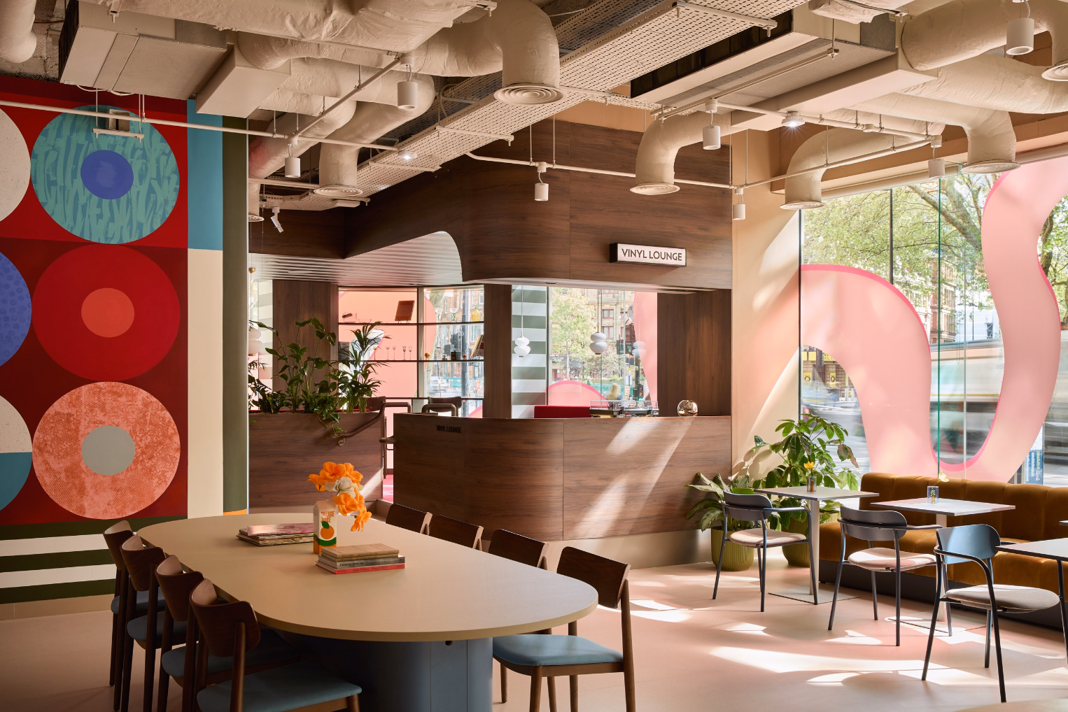 Brown chairs sit at a cream table across from black chairs, grey tables, and a yellow sofa. A wood counter stands under white pipes next to a circle mural.