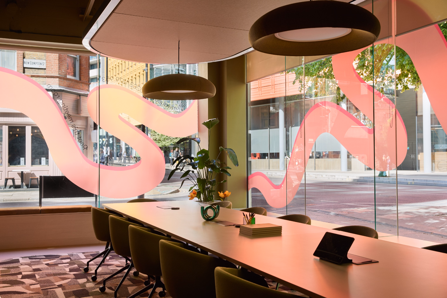 Green chairs sit at a cream table on a rug. Green lights hang from a ceiling near a plant and windows with a pink graphic.