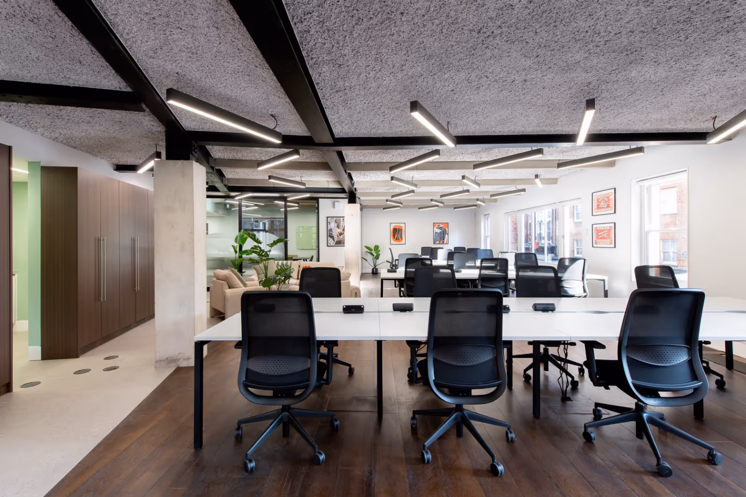 Black chairs at white desks on a wood floor near wood cabinets and a cream sofa. White lights and black beams are on a grey ceiling.