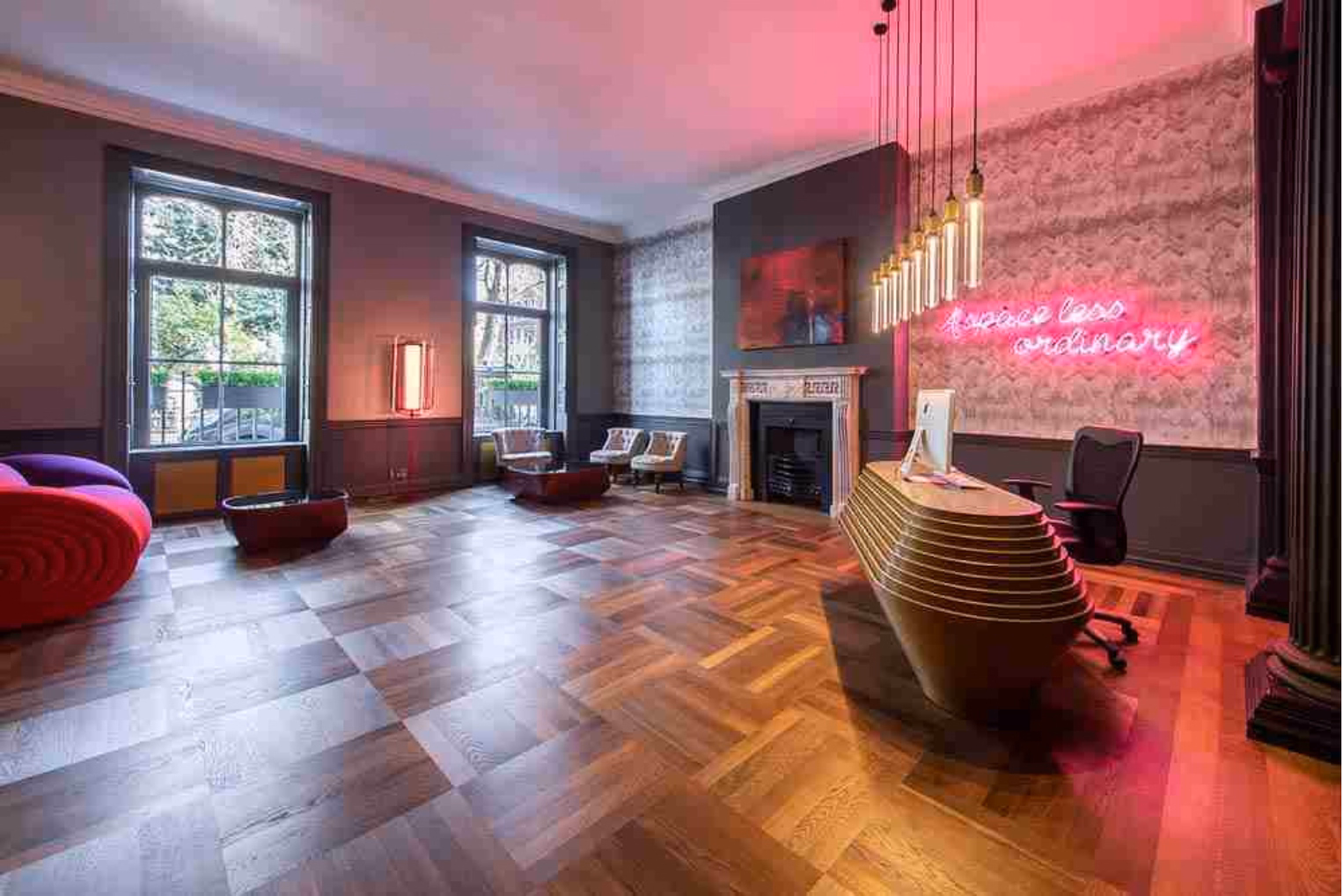  A gold desk, a black chair, a red and purple seat, and red tables on the wood floor. A pink neon sign hangs on a wall near a fireplace and windows.