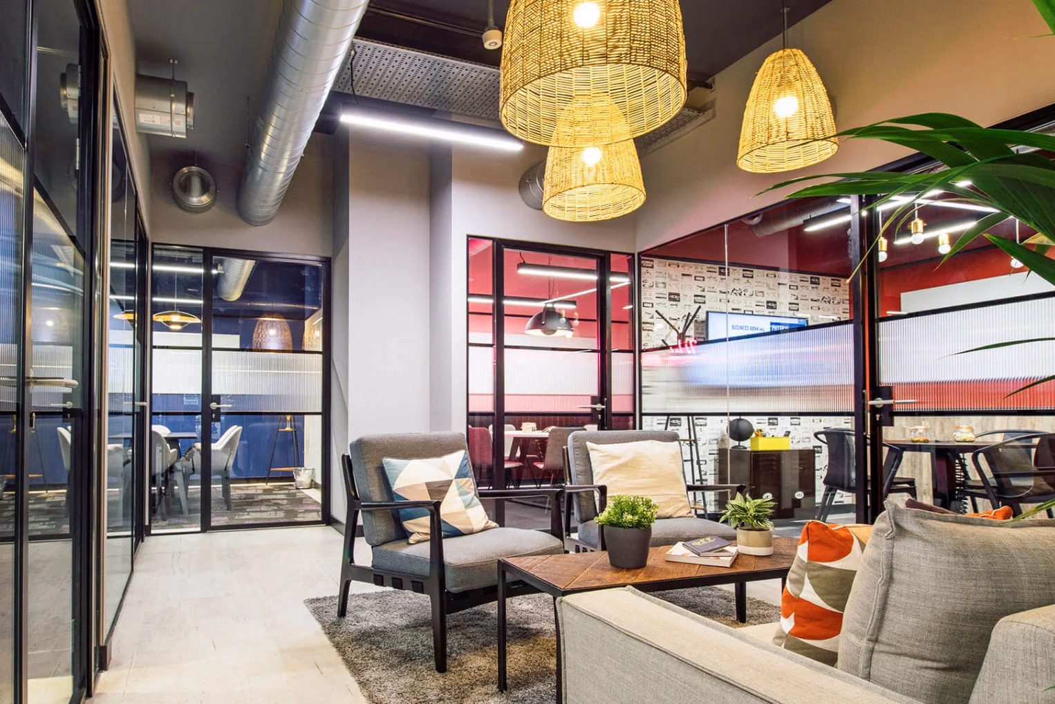 Two grey armchairs and a grey sofa near a wood table on a grey rug. Woven lights and a silver pipe hang from a ceiling next to glass rooms with black frames.