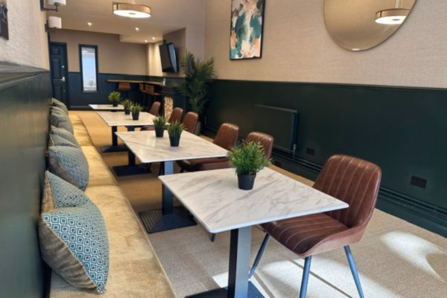 Brown chairs and a cream bench with blue pillows at white marble tables. Small plants on the tables in a room with green walls and a round mirror.