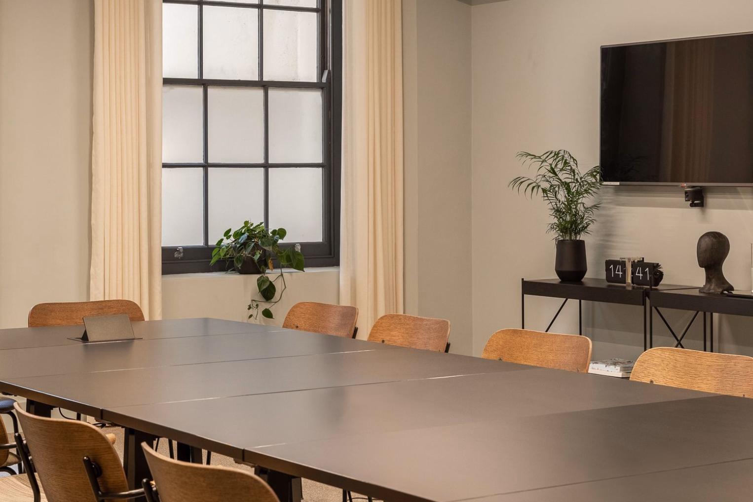 Brown chairs sit at a grey table near a black console table. Plants, a clock, and a TV are in a room with a window and white curtains.