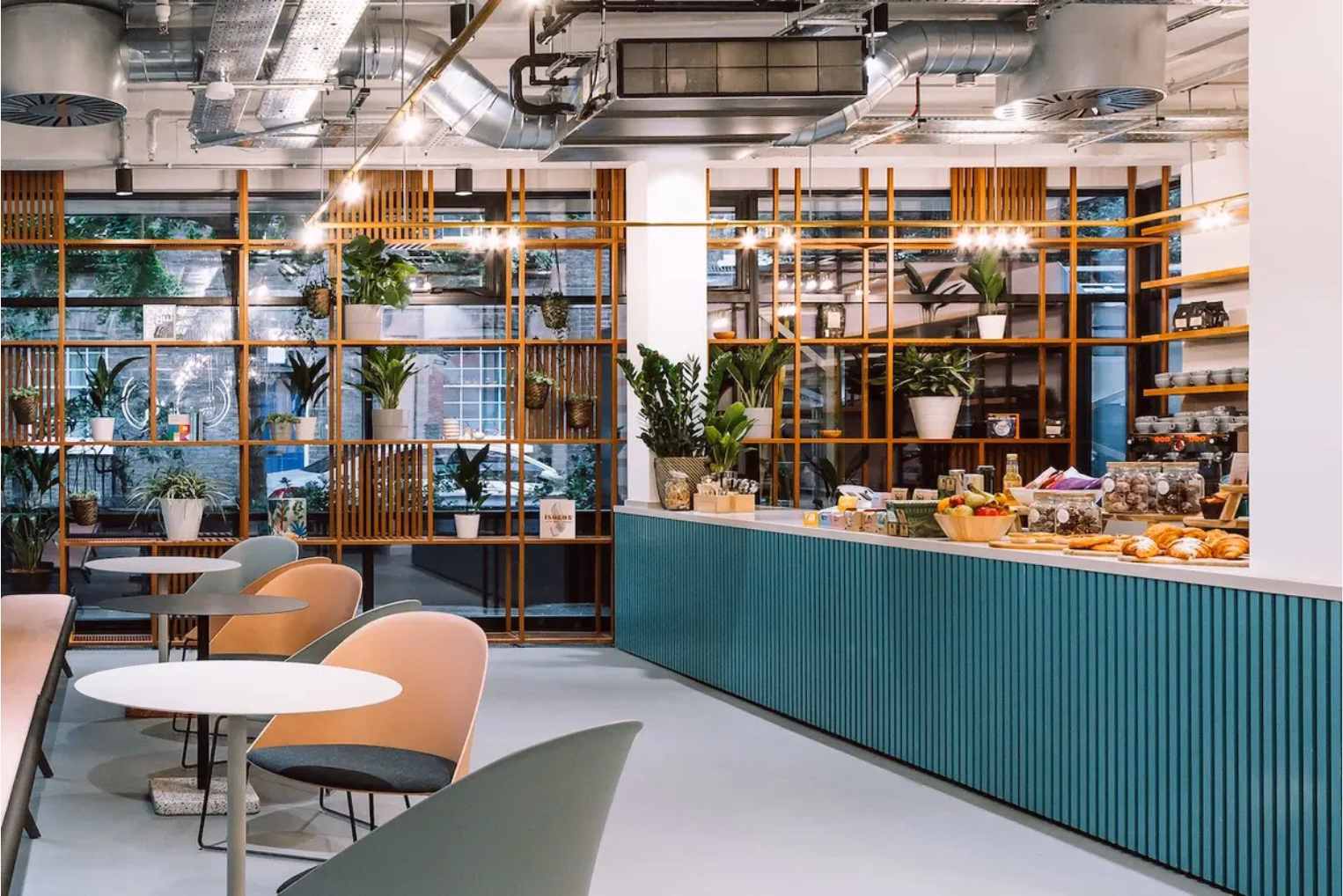 Pink and grey chairs at white and grey tables near a teal counter. Plants sit on wood shelves under silver pipes on a ceiling.