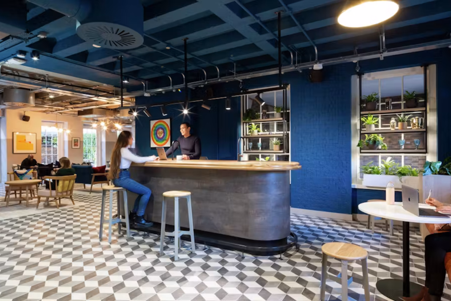 A woman and a man talk at a grey and wood counter. A person uses a laptop at a white table, and wood stools sit on a grey and white floor under a blue ceiling.