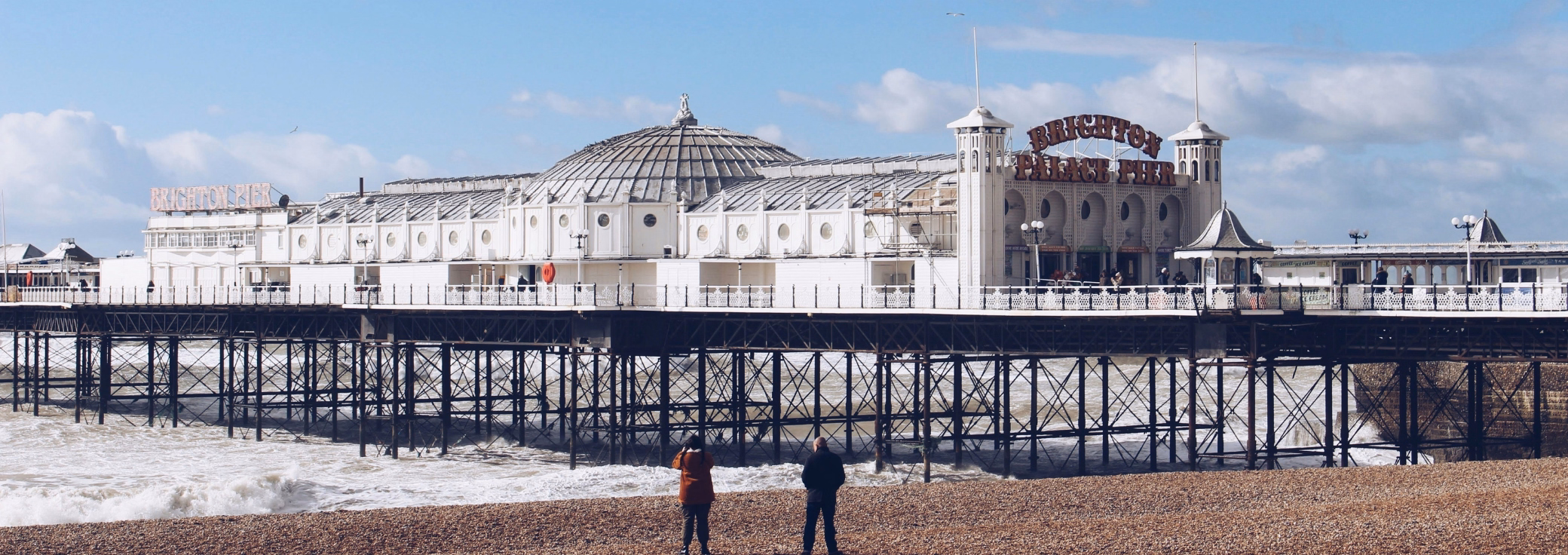 Two people stand on a beach looking at a white pier over the sea under a blue sky with clouds.