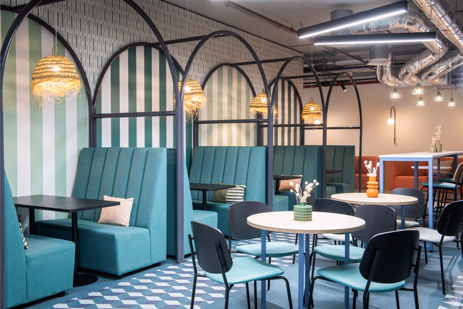 Teal booths and black tables stand under black arches next to a striped wall. Teal and black chairs sit at wooden tables on a checkered floor under ceiling pipes.