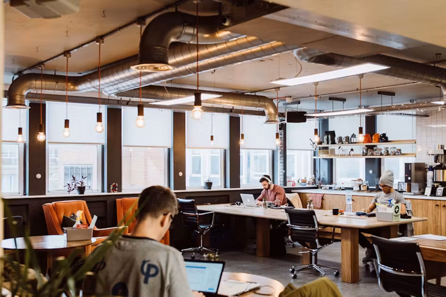 People use laptops at wooden tables with black chairs and orange armchairs. Lights and silver pipes hang from the ceiling.