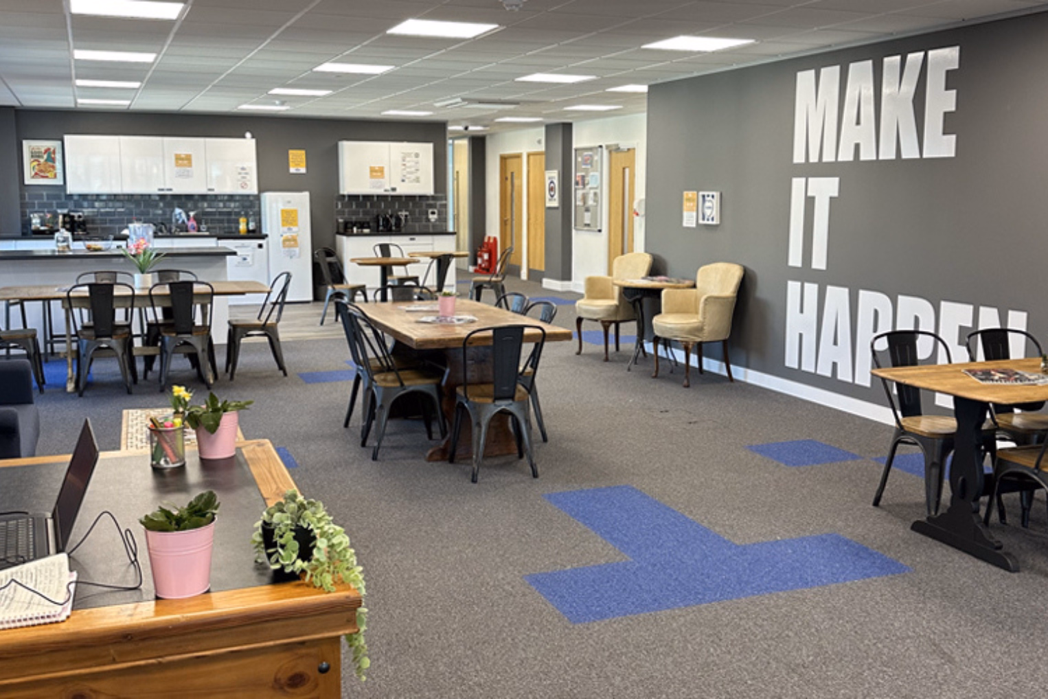 Black chairs sit at brown tables on a grey and blue floor. Cream armchairs are near a grey wall with text in front of white kitchen cabinets.