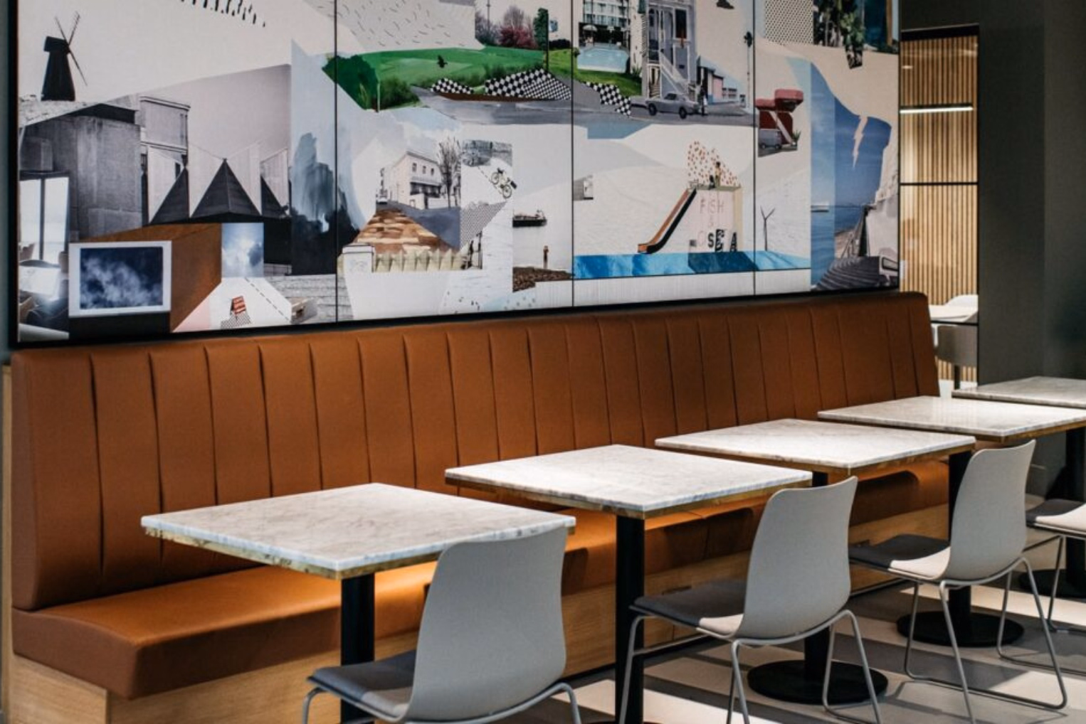 Grey chairs sit at white marble tables in front of a brown booth under a collage mural on a wall.