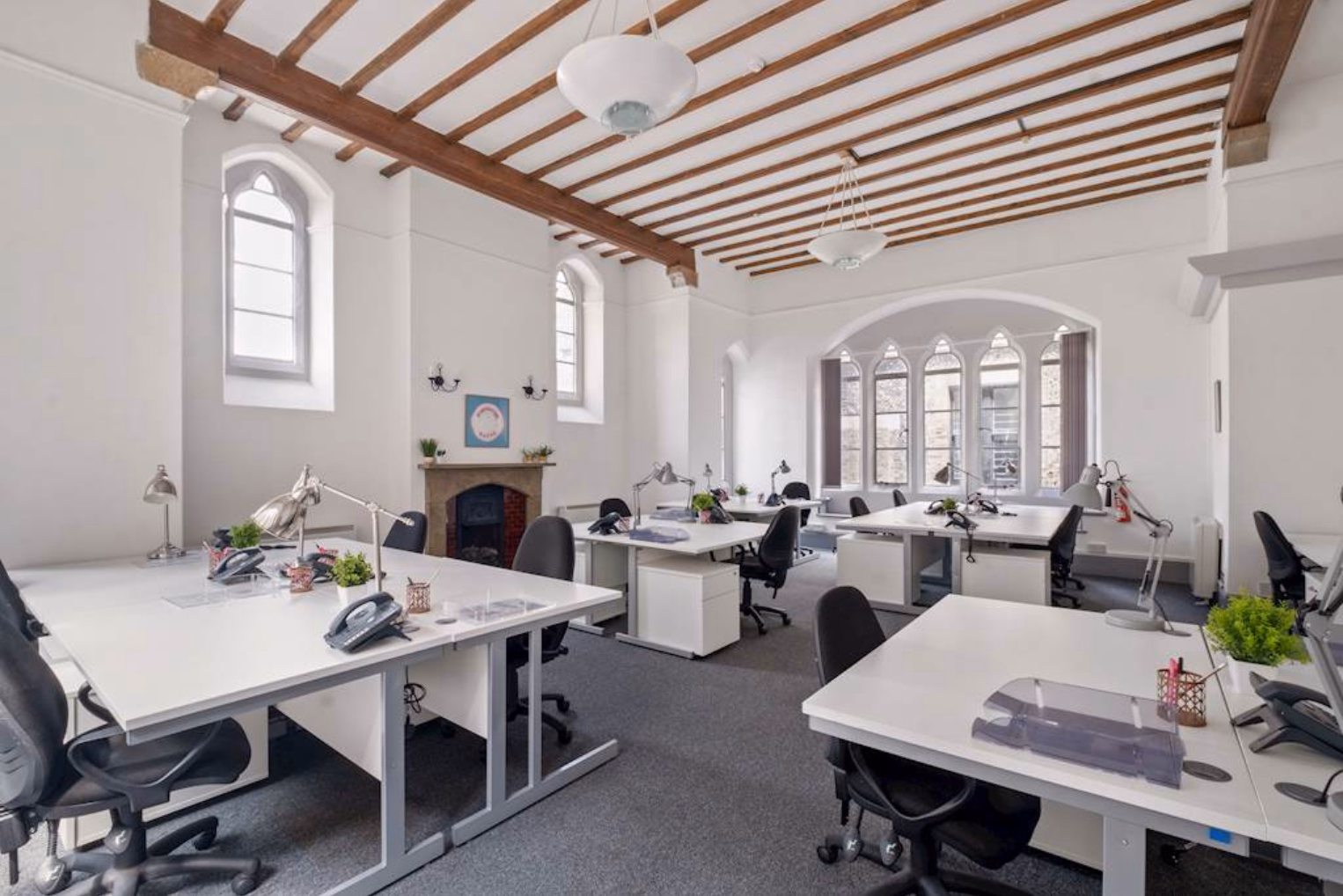 Black chairs sit at white desks on a grey floor. Wood beams and white lights are on the ceiling near a fireplace and windows.