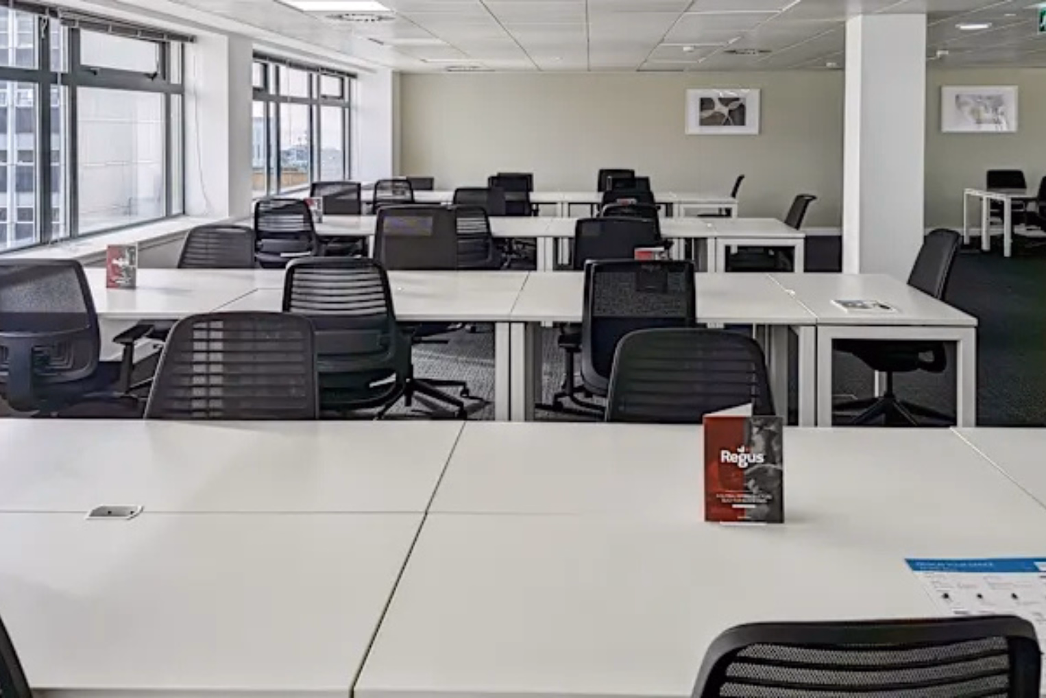 Black chairs sit at white desks on a grey floor near windows and white pillars under a tiled ceiling.